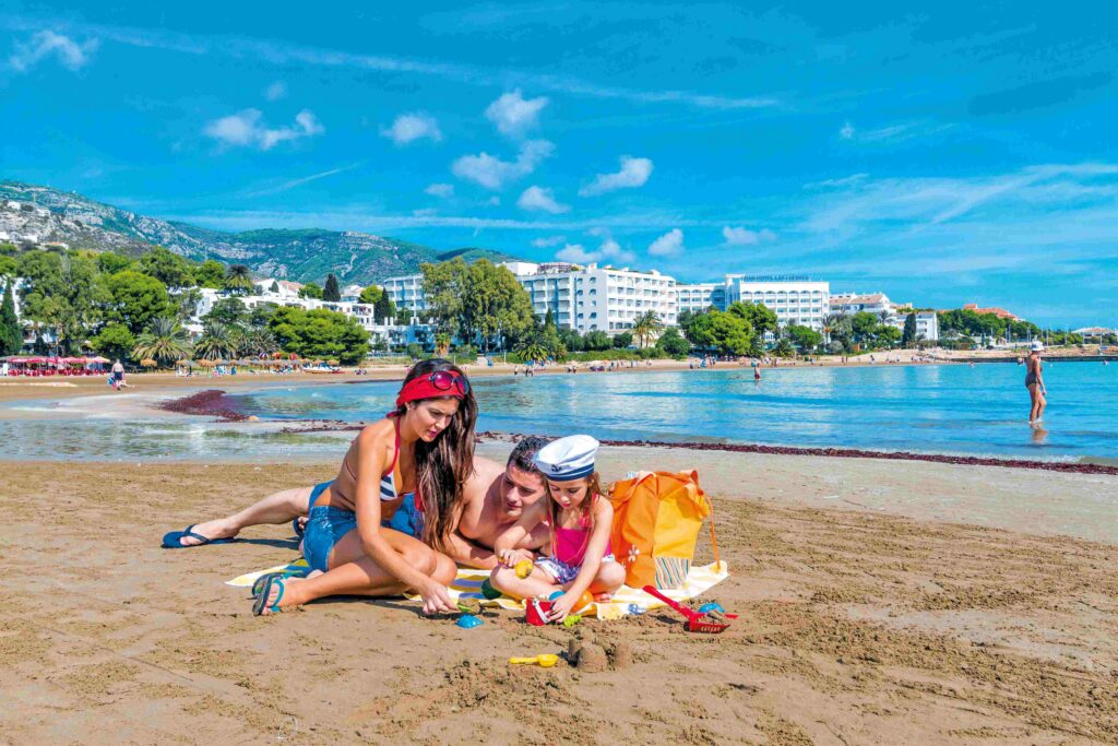 Woman and two children building sandcastles on a sunny beach with resort buildings and hills in the distance.