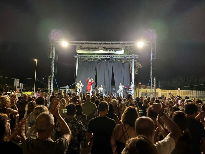 Crowd at an outdoor night concert, performers on a lit stage with a banner overhead and illuminated by bright spotlights.
