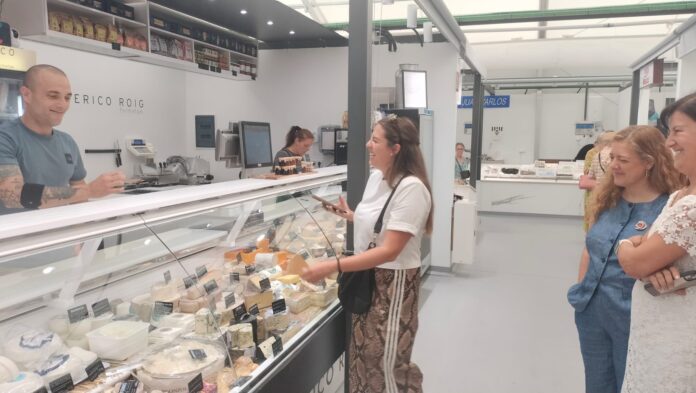 Deli counter with a smiling male staff member serving two women customers, cheese blocks and wheels displayed in a glass case.