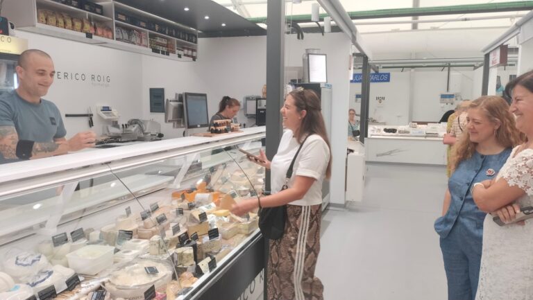 Deli counter with a smiling male staff member serving two women customers, cheese blocks and wheels displayed in a glass case.