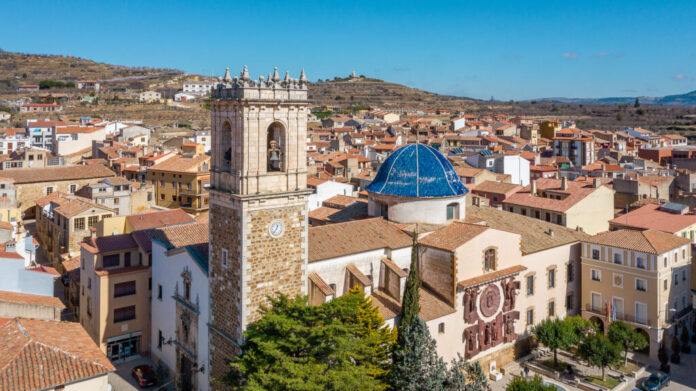 Aerial view of a sunny Mediterranean town with a stone clock tower and a blue-domed church among terracotta rooftops.
