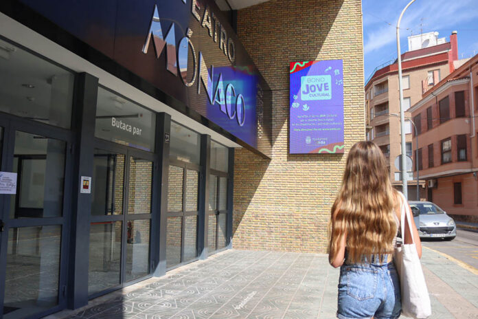 Young woman with long wavy hair in denim shorts walks along a storefront with glass doors and a bright blue sign above.