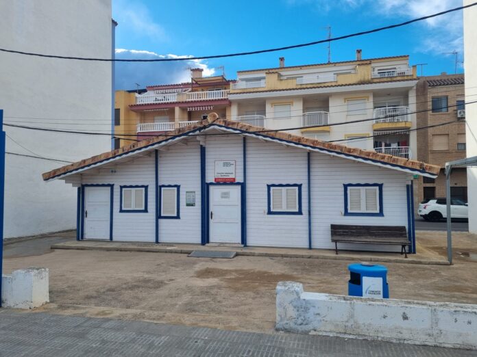 Blue-and-white wooden storefront with closed shutters and a bench in front, residential buildings behind under a bright blue sky with power lines overhead.