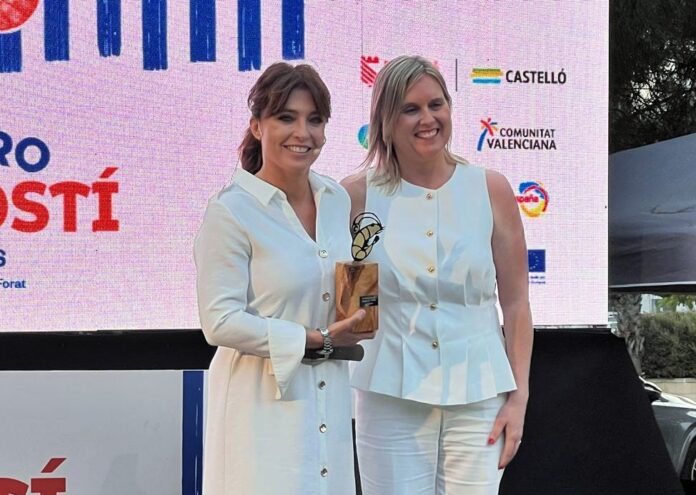 Two women in white outfits smile on stage as one holds a wooden trophy with a metal abstract sculpture, posing for a photo with a pink sponsor backdrop behind them.