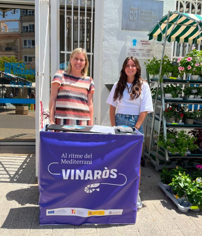 Two smiling women stand behind a purple market stall banner that reads VINARÒS outside a flower shop setting.