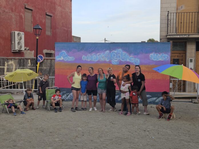 Group of adults and children posing for a photo in front of a colorful mural on a sunny outdoor set with umbrellas nearby.