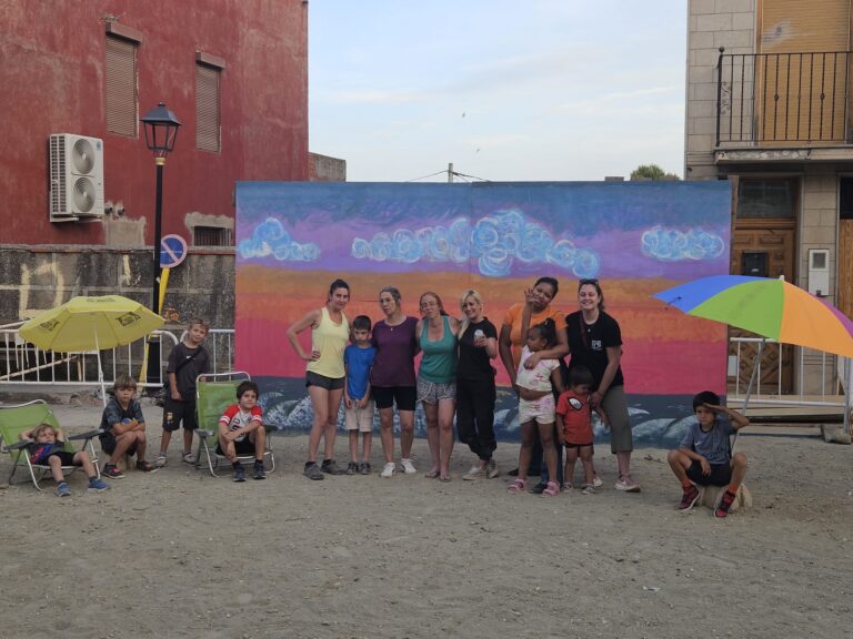 Group of adults and children posing for a photo in front of a colorful mural on a sunny outdoor set with umbrellas nearby.