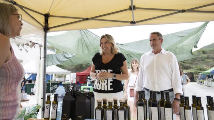 People at an outdoor market stall with bottles of oil on the table under a yellow canopy.