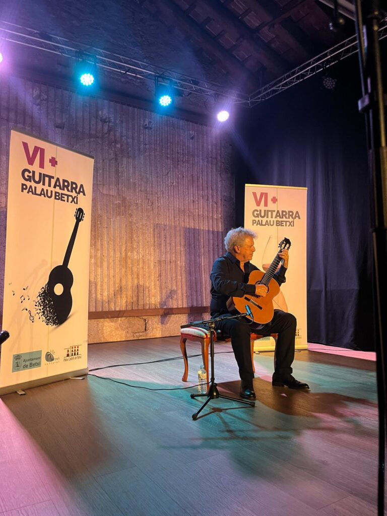 Male guitarist seated on stage, playing a classical guitar at a VI+ Guitarra Palau Betxi event with banners beside him.