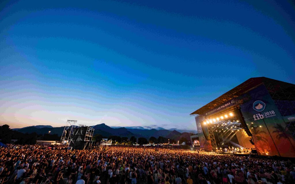 Large outdoor concert at dusk with a packed crowd and a bright stage on the right against a blue sky.