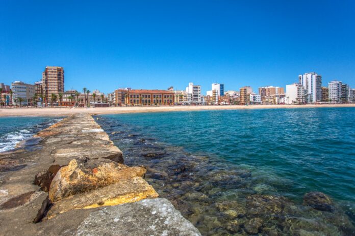 Stone breakwater extending into turquoise sea toward a sandy waterfront with a row of low- to mid-rise buildings under a clear blue sky on a sunny day.
