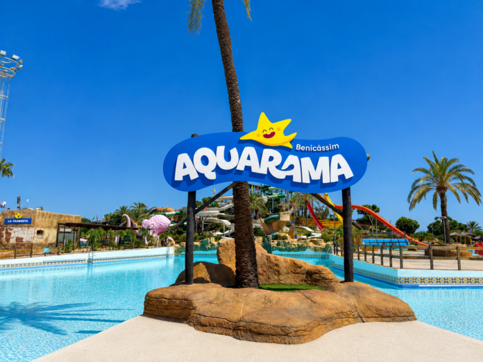 Aquarama water park sign on a rocky island, with a smiling yellow sun graphic above the blue logo under a clear sky.