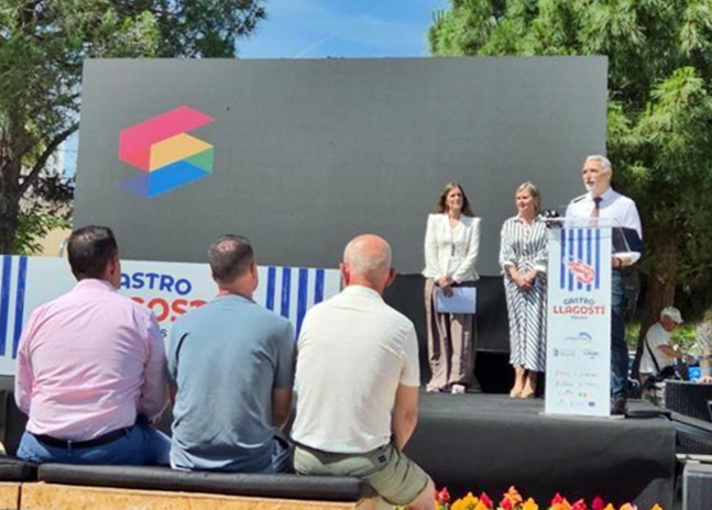 Outdoor stage with three speakers at a podium, discussing a bannered event under a large backdrop and trees in the background.