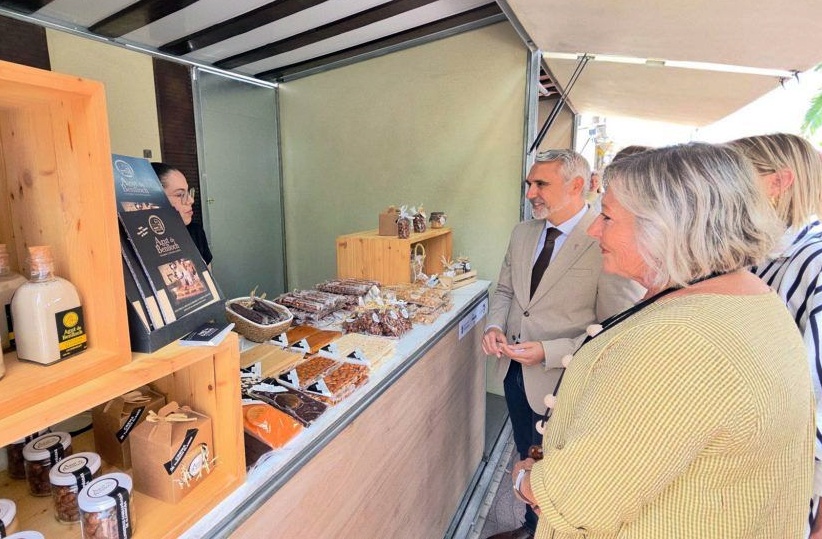 People gathered at an outdoor market stall inspecting a display of packaged foods and jars on the counter.