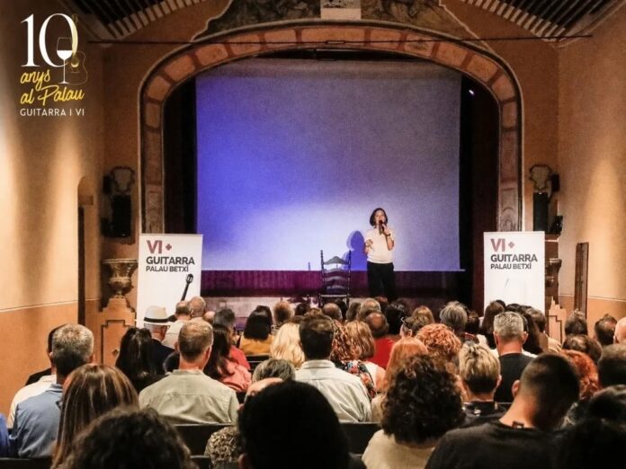 Singer performs on a small stage in a historic theater, with a purple backdrop and a seated audience watching. Banners read 'VI+ GUITARRA PALAU BETXI'.