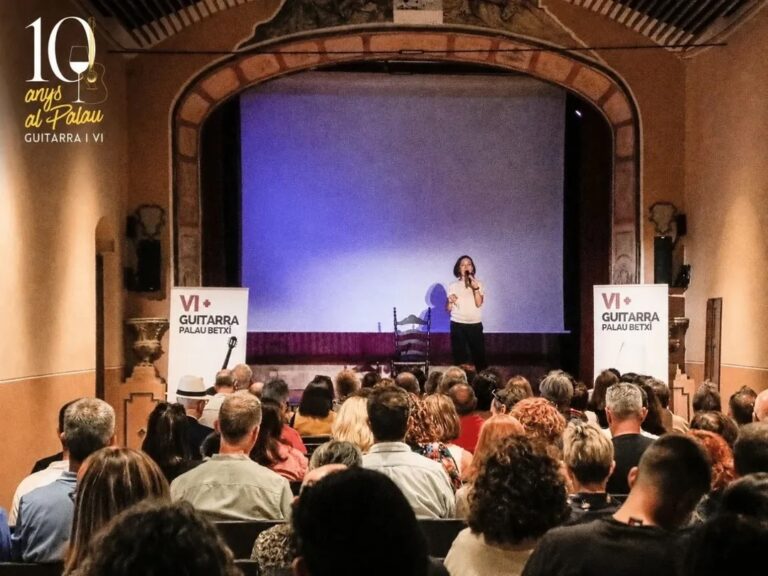 Singer performs on a small stage in a historic theater, with a purple backdrop and a seated audience watching. Banners read 'VI+ GUITARRA PALAU BETXI'.