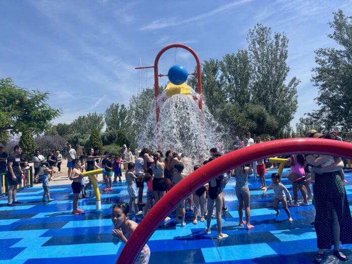 Children playing in a splash pad with a blue globe sculpture under a red arch and water spraying everywhere.