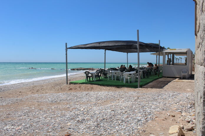 Beach cafe on a pebble beach with a black shade canopy and white plastic chairs facing turquoise sea under a clear blue sky.