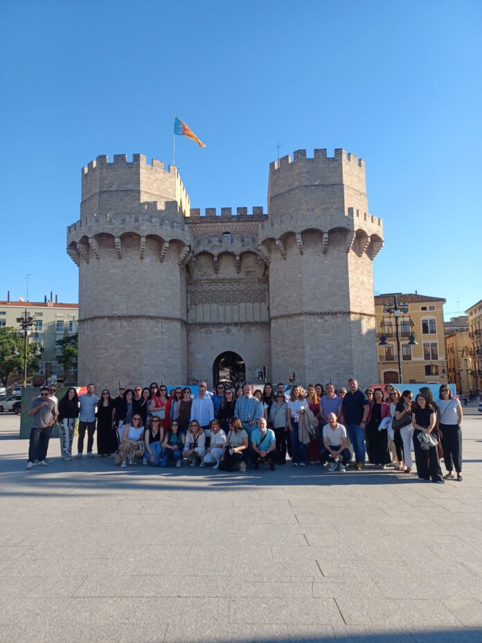 Group of people posing for a photo in front of a historic stone fortress with two towers under a blue sky (flag on left tower).