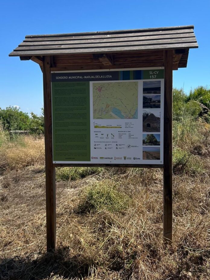 Outdoor wooden information board with a roof, displaying a map and photos, standing in a dry grassy area under a clear blue sky.