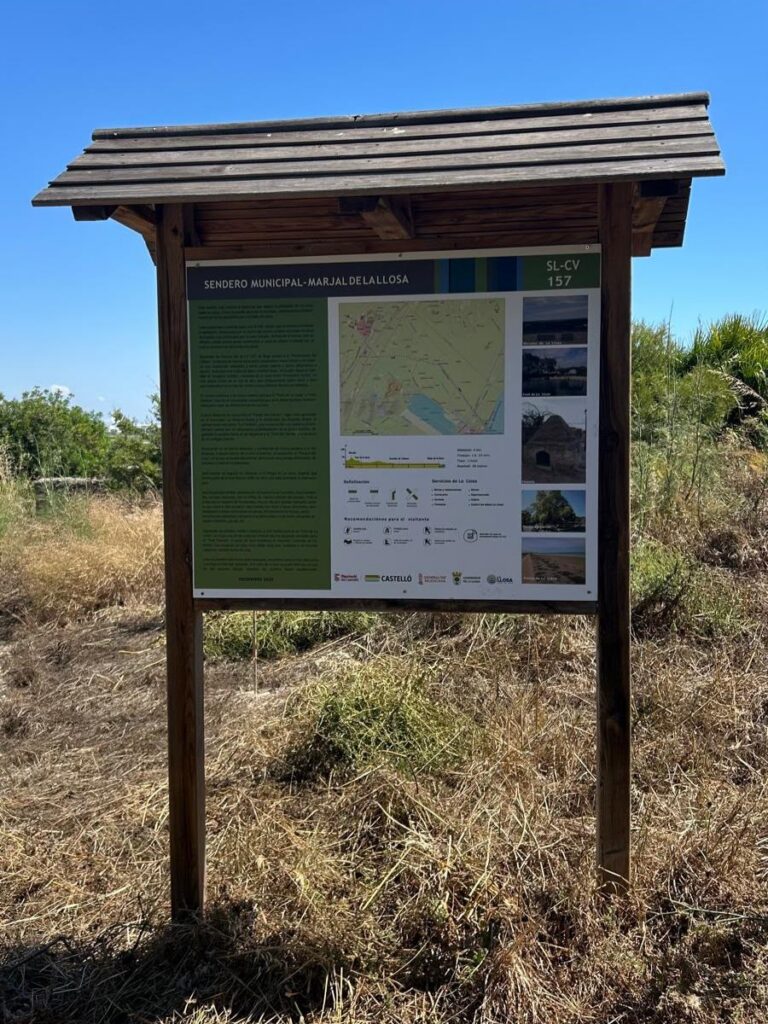 Outdoor wooden information board with a roof, displaying a map and photos, standing in a dry grassy area under a clear blue sky.