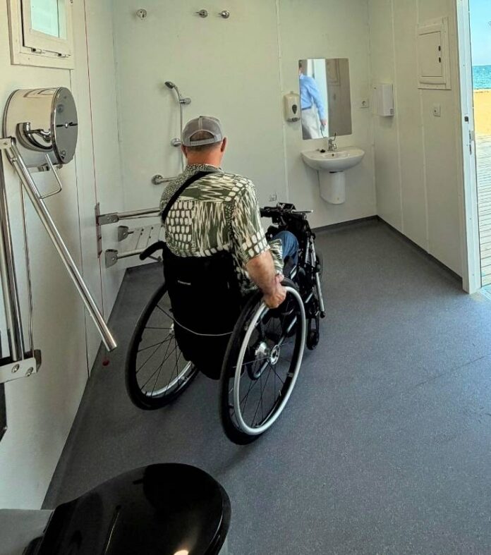 Person in a wheelchair inside an accessible bathroom with grab bars, a wall-mounted sink, and an open door to an outdoor deck.