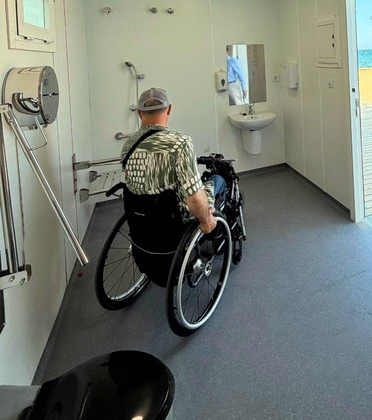 Person in a wheelchair inside an accessible bathroom with grab bars, a wall-mounted sink, and an open door to an outdoor deck.