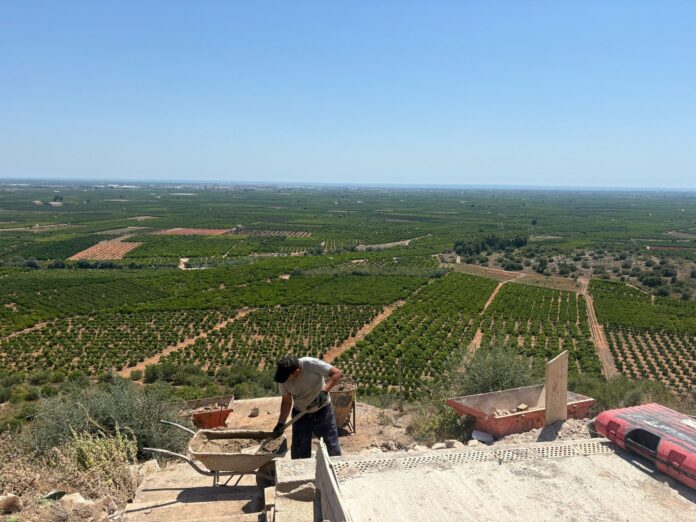 Construction worker pushing a wheelbarrow filled with concrete at a dusty building site, with patchwork fields and a distant town on the horizon under a clear blue sky