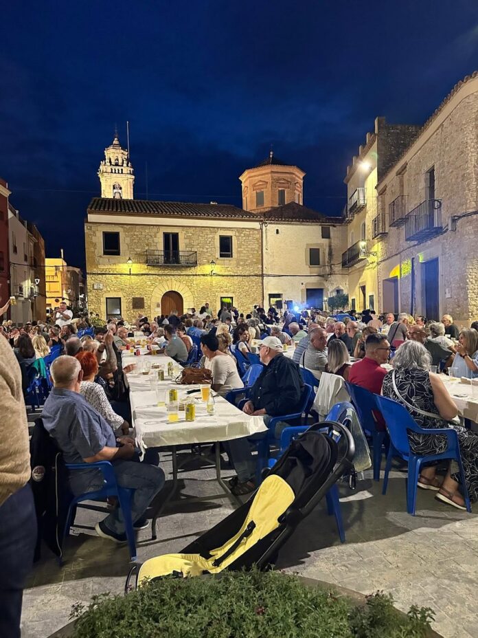 Nighttime outdoor communal dinner in a historic town square with long tables and many guests seated on blue chairs under warm lights.