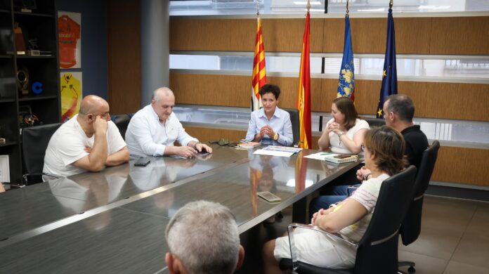 Group of professionals seated around a long conference table in a meeting room, with several flags displayed in the background while they discuss documents.