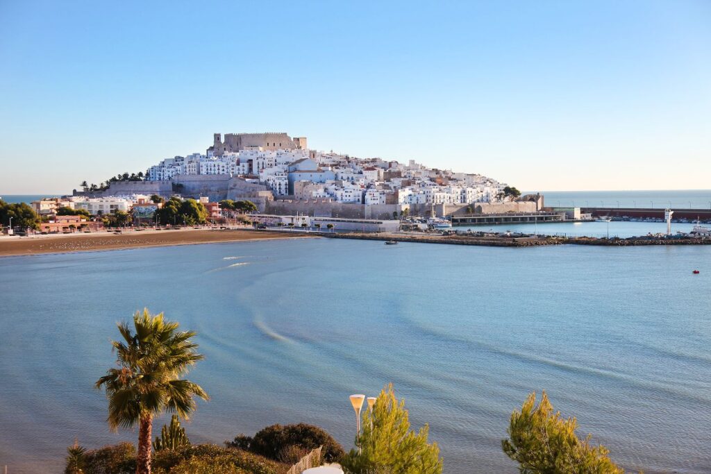 White-washed hillside town with a medieval fortress, overlooking a calm blue bay; palm trees in the foreground.