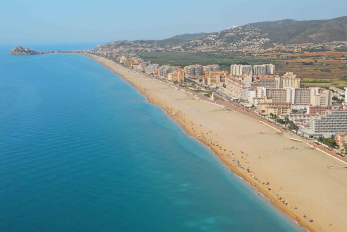 Aerial view of a long sandy beach, turquoise sea, and a city of high-rise buildings along the coast with hills in the background.