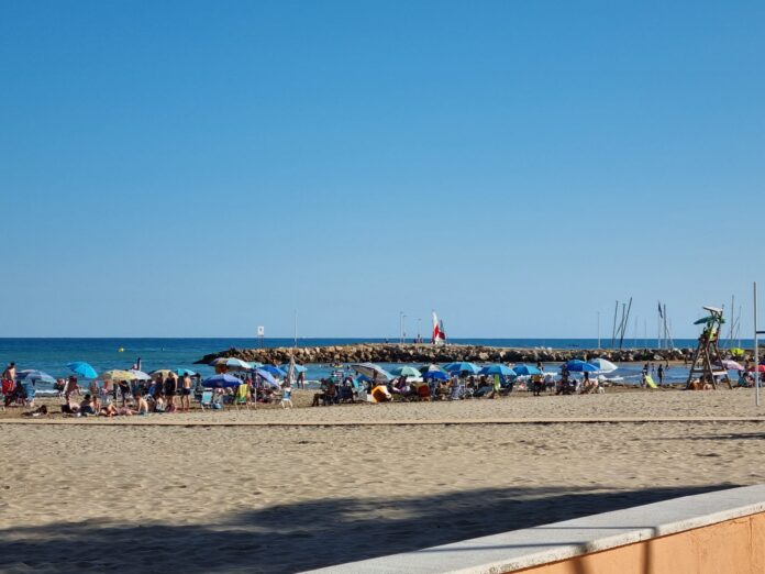 Crowded sandy beach with many colorful umbrellas and sunbathers, a rocky breakwater stretching into the blue sea under a clear sky, lifeguard tower on the right.