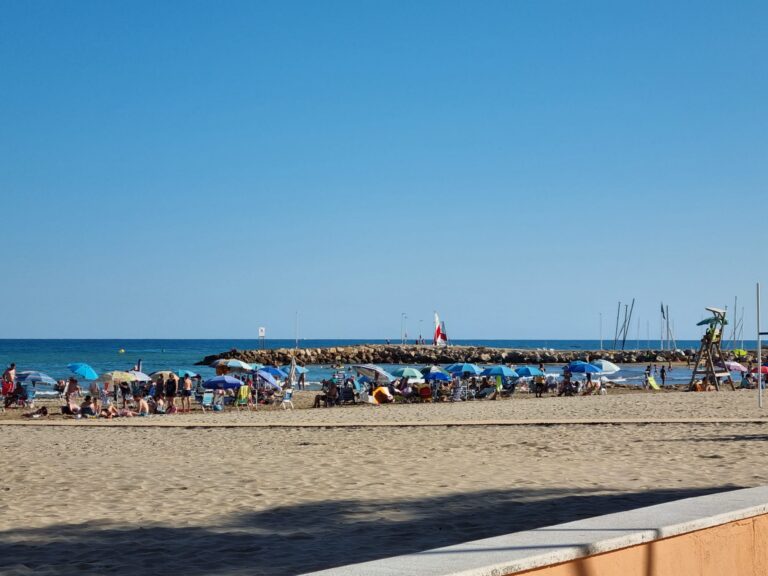 Crowded sandy beach with many colorful umbrellas and sunbathers, a rocky breakwater stretching into the blue sea under a clear sky, lifeguard tower on the right.