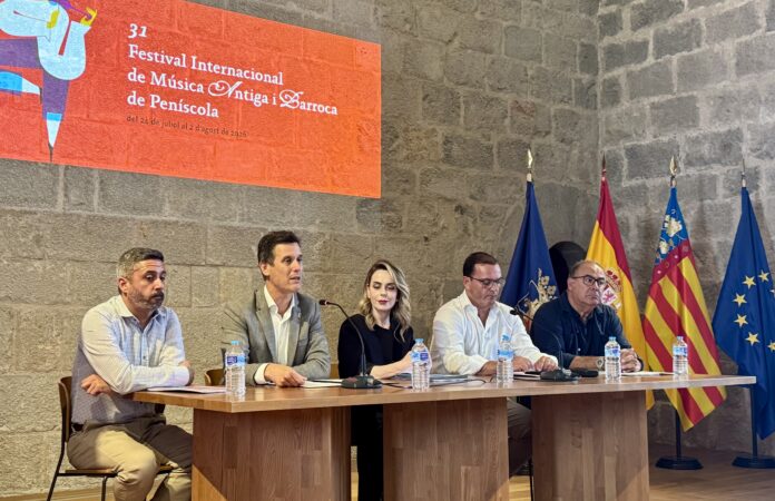 Panel of six people seated at a wooden table in a stone-walled room, with an orange festival banner and multiple flags behind them (Spain and EU flags visible). They have microphones and water bottles on the table, suggesting a press conference or panel discussion at a cultural festival.
