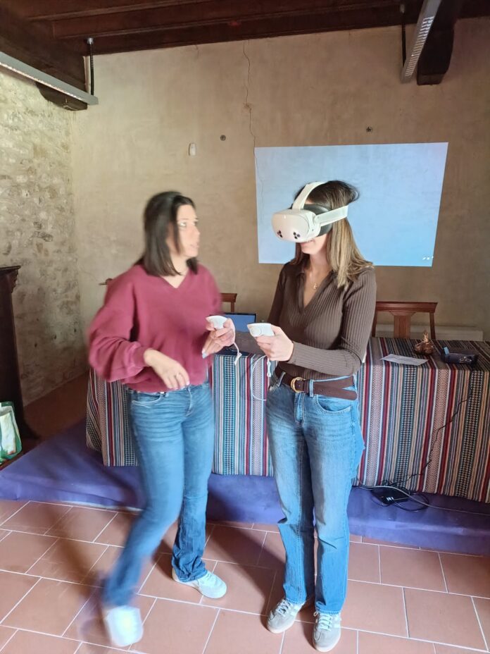 Two women indoors; one wears a white VR headset while the other chats and holds a small device or controllers.