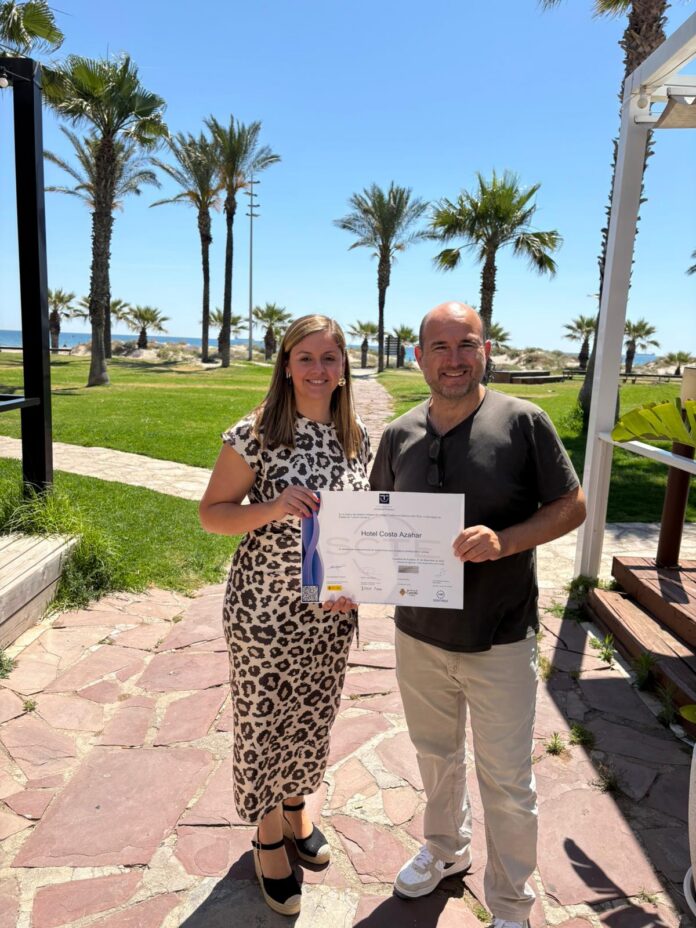 Two adults stand on a sunny seaside path holding a certificate, with palm trees and the ocean in the background.