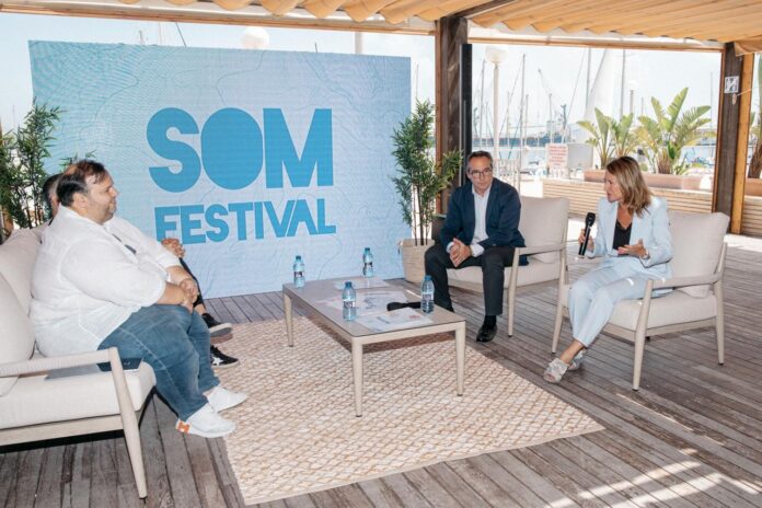 Host with microphone interviews two guests on a marina deck during SOM Festival; water and boats visible in the background.