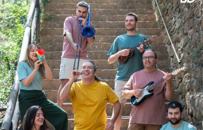 A group of seven adults on outdoor stone stairs playing music together, with guitars, a ukulele, a trumpet, and a woman blowing a red toy horn.