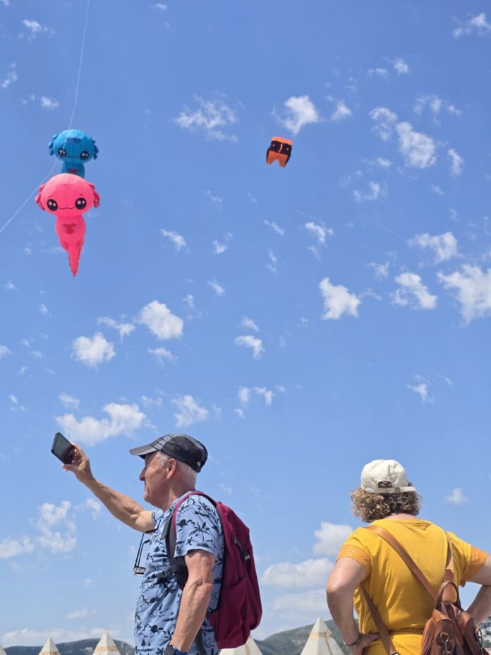 Two people, one taking a selfie, watch large pink and blue cartoon kites float in a clear blue sky with small orange kite nearby.
