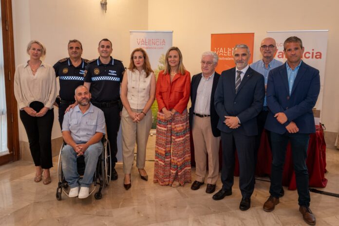 A group of ten people posing for a formal photo indoors at an event, including two police officers in uniform and a man in a wheelchair, with Valencia banners in the background.