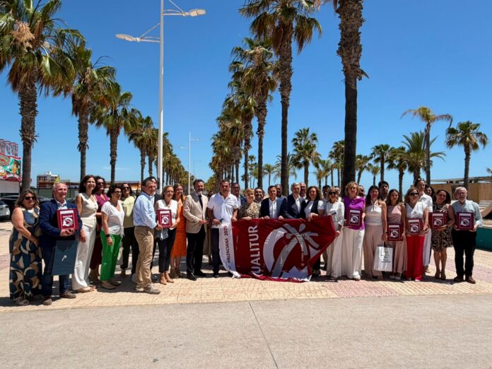 Group of about 25 adults posing for a photo outdoors on a sunny day, holding red award plaques and a large maroon banner with a white logo, palm trees lining a promenade behind them.