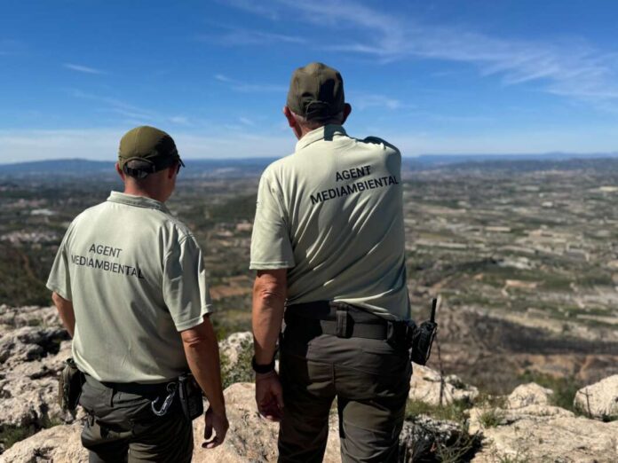 Two uniformed agents stand on a rocky cliff, backs to the camera, overlooking a valley and distant hills under a clear blue sky.