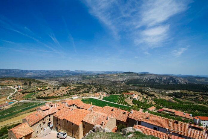 Cluster of stone houses with red-tiled roofs along a hillside edge, overlooking green fields and rolling hills under a bright blue sky.