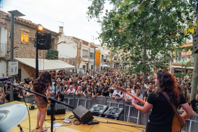 Crowd of people watching a live outdoor concert on a street stage with musicians and trees overhead.