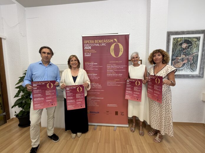 Four adults stand indoors, holding pink event flyers in front of a tall promotional banner for Opera Benicàssim 2026.