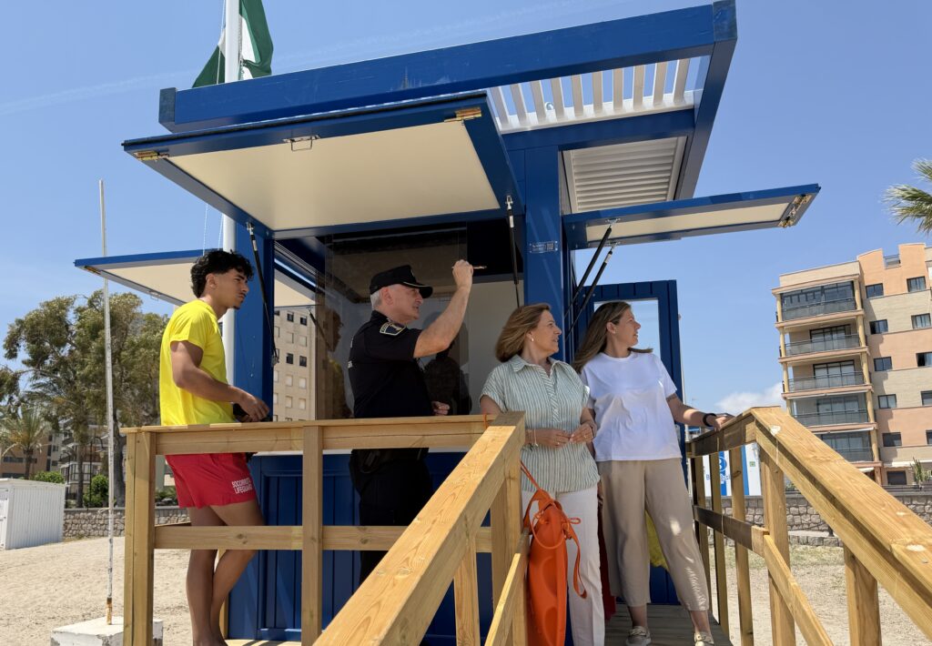People stand on a blue lifeguard station platform with a wooden railing, looking outward on a sunny day.