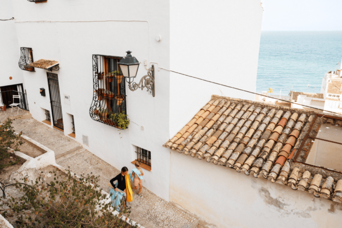 Two women walk a cobblestone path beside white houses with iron-window grilles, near the sea.
