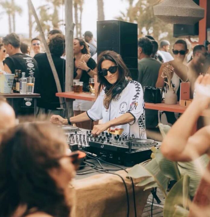 Female DJ wearing sunglasses mixes tracks at an outdoor party with a crowd around a palm-tree setting.