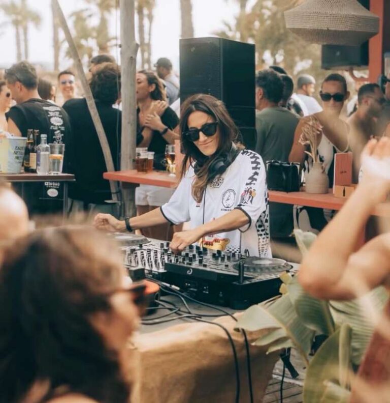 Female DJ wearing sunglasses mixes tracks at an outdoor party with a crowd around a palm-tree setting.